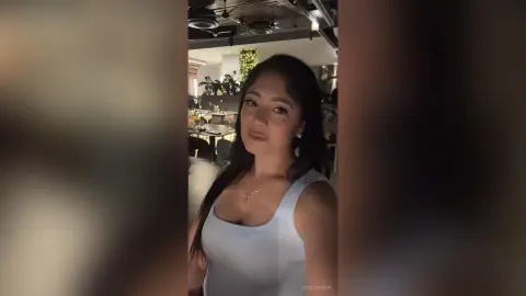 Smiling young woman in a white tank top posing in a modern, dimly lit restaurant setting.