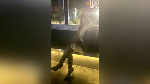 Elegant woman in a black sequin top and tall leather boots sitting in a dimly lit lounge at night.
