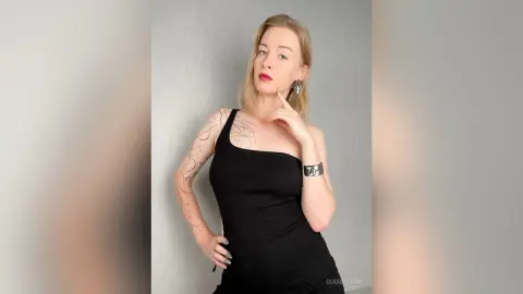Elegant woman in a black one-shoulder dress and silver jewelry posing against a grey wall.