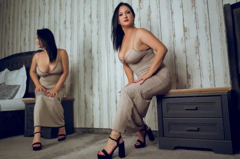 Elegant woman in a beige midi dress posing against a rustic wood wall in a moody indoor setting.