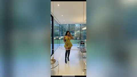 Stylish woman in a yellow mini dress posing in a modern, bright luxury apartment with large glass windows.