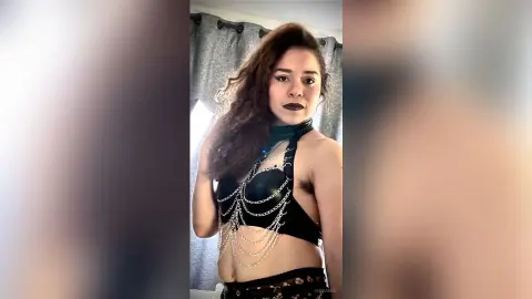 Young woman in a black lace bra and silver chain harness posing confidently in a bright room.