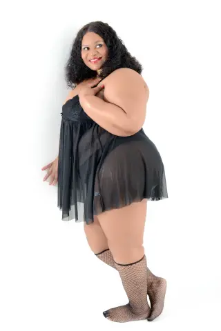 Curvy woman in a black sheer nightgown and fishnet stockings posing against a white background.