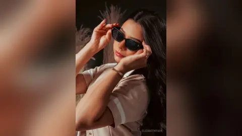 Stylish woman in a beige short-sleeved shirt wearing black sunglasses in a warm, moody indoor setting.