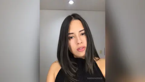 Young woman with long dark hair wearing a black sleeveless top in a minimalist indoor setting.