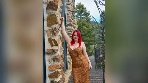 Red-haired woman in a glamorous gold evening gown posing by a rustic stone wall in a scenic outdoor setting.