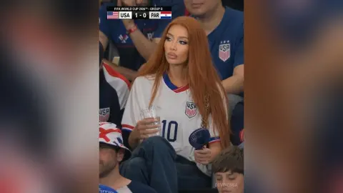 DeviNessa wears a white USA soccer jersey and holds a clear glass while sitting in the stands.