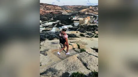 AnaMathers stands on a rocky cliffside overlooking a coastal town, wearing a white sleeveless top and dark shorts.