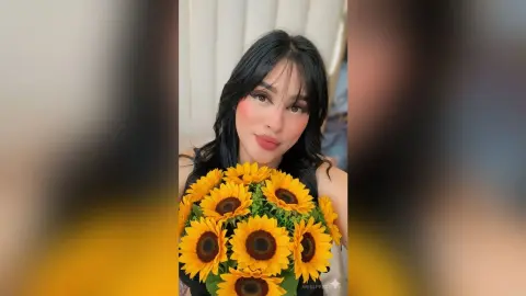 Smiling young woman with long dark hair holding a bright bouquet of sunflowers in a soft indoor setting.