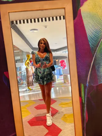 Woman in a green floral mini dress taking a mirror selfie in a modern, brightly lit hotel lobby.