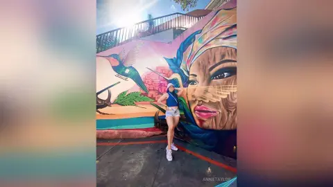Young woman in a blue tank top and white shorts standing before a colorful large-scale street art mural.