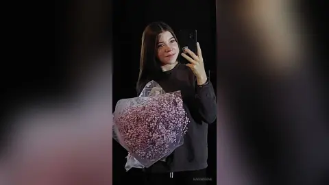 Smiling young woman in a black long-sleeve shirt holding a large bouquet of pink flowers in a dark setting.
