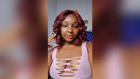 JenniferJoe poses in a light pink lace-up tank top that shows her cleavage, looking directly at the camera with wavy red hair.