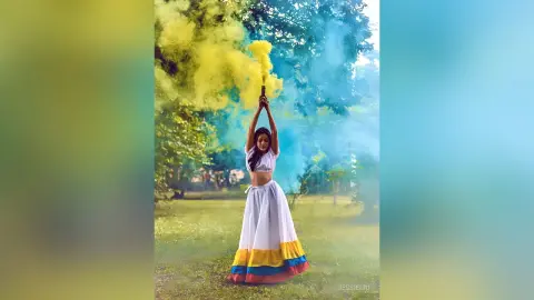 JessieLiu stands in a grassy field holding a yellow smoke flare above her head, wearing a white crop top and a long white skirt with yellow, blue, and red stripes.