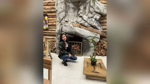 Smiling woman in a black leather jacket and jeans sitting in a rustic log cabin living room.