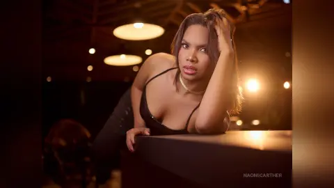 Glamorous woman in a black sleeveless top leaning on a bar in a warm, dimly lit lounge setting.