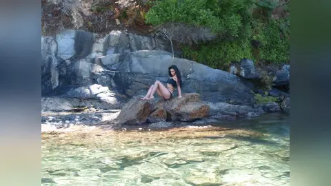 Woman in black bikini relaxing on rocky shore by clear turquoise ocean water under bright sunlight.