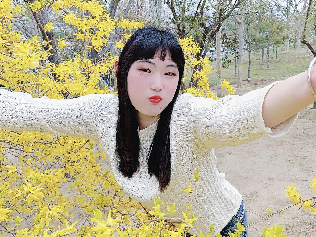 LouiseAnna poses outdoors against a backdrop of bright yellow flowering bushes, wearing a white long-sleeved knit top.