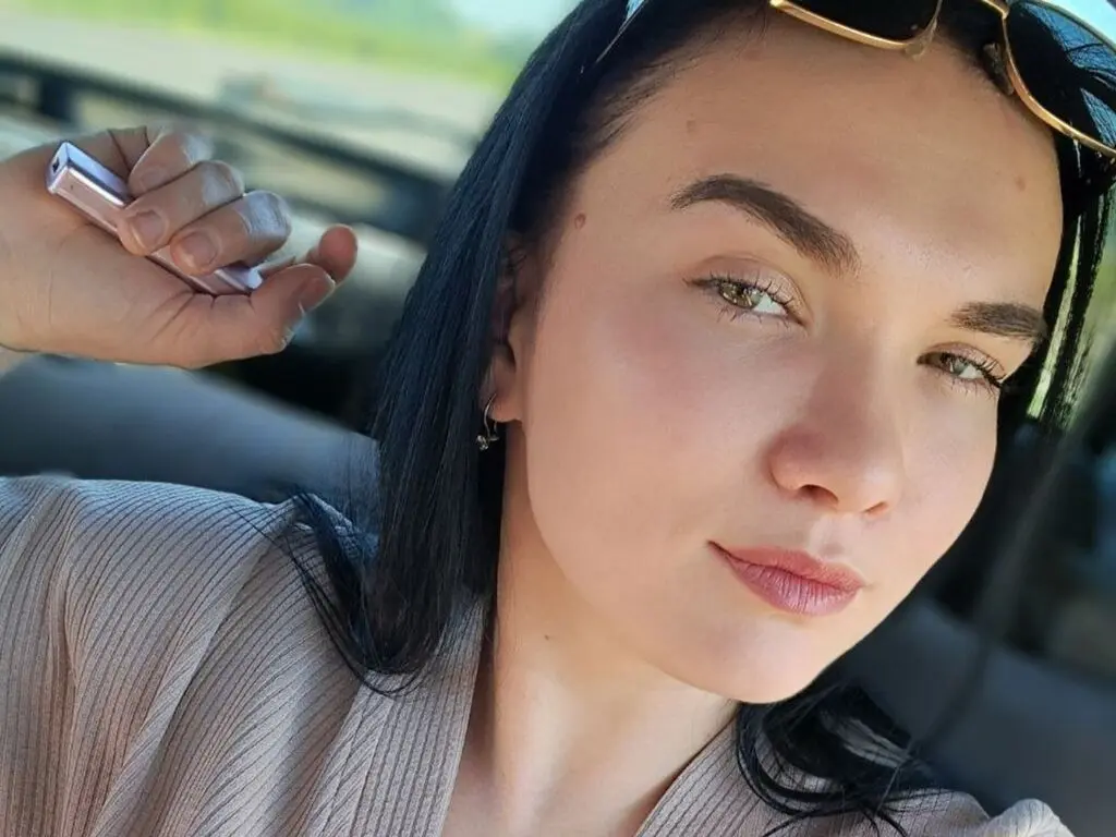 Smiling woman in a beige ribbed top sitting in a car with sunglasses on her head during a sunny day.