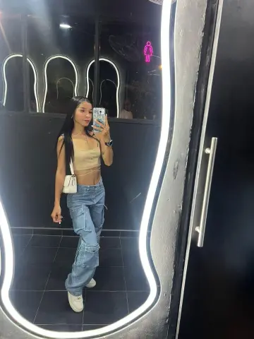 Young woman in a beige crop top and baggy blue jeans taking a mirror selfie in a modern restroom.