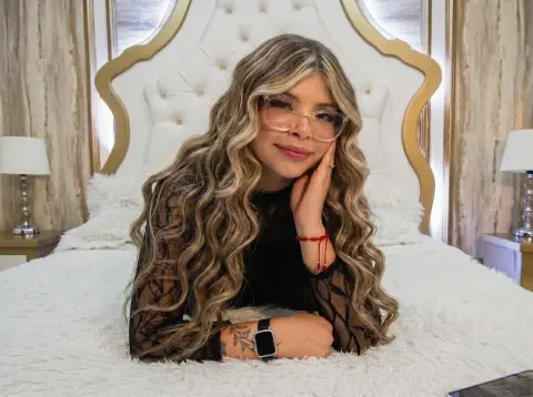 RiileyAngel with long wavy blonde hair and glasses lies on a white bed wearing a black sheer lace top.