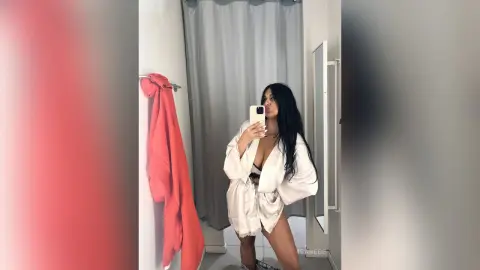 Young woman in a white satin robe taking a mirror selfie in a bright, minimalist modern bathroom.