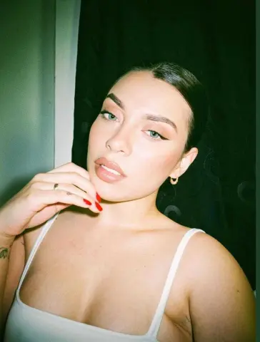 Young woman with sleek hair and winged eyeliner wearing a white tank top in a moody indoor setting.