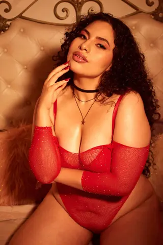 Curvy woman in red mesh bodysuit sitting on a bed with a glamorous, romantic mood.