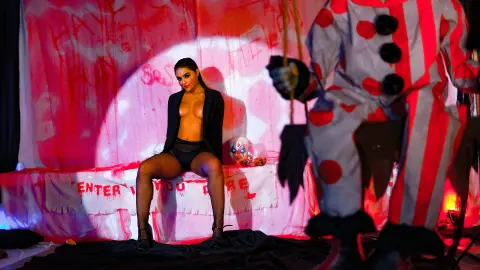Glamorous woman in black lingerie sitting on a stage under pink spotlight with a clown performer in the foreground.