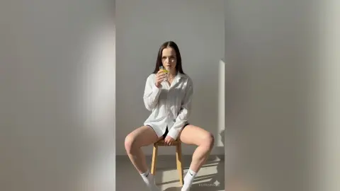 Young woman in a white button-down shirt sitting on a wooden stool in a bright, minimalist room.