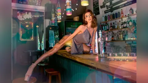Glamorous woman in a sparkling silver dress posing on a wooden bar in a moody, dimly lit nightclub setting.