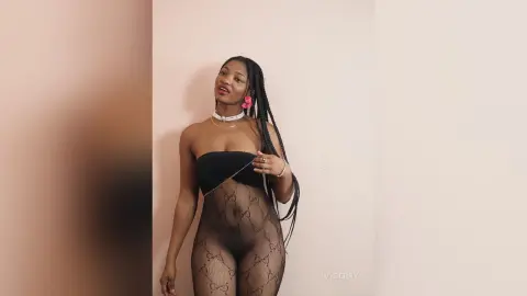 IvyBerry poses in a black tube top and patterned black fishnet tights against a plain wall, showing her long dark braids and white choker.