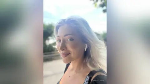 Smiling blonde woman in a black tank top wearing wireless earbuds outdoors in soft natural light.