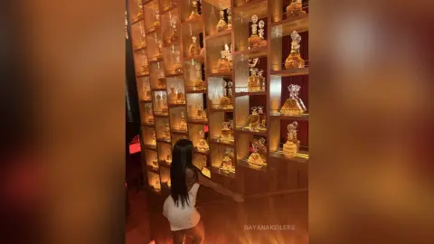 Woman in a white dress browsing luxury perfume bottles on a glowing wooden display shelf in a warm boutique.