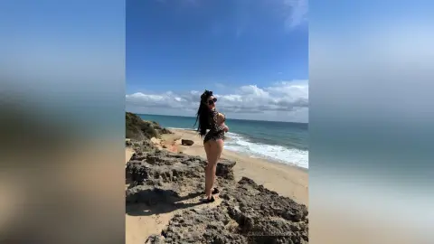 Stylish woman in a black patterned swimsuit posing on a sunny rocky beach by the blue ocean.