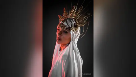 LeslyAnderson wears a gold crown and white silk headwrap against a dark background with red lighting.