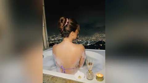 LaraCole shows her bare back and shoulders while relaxing in a bubble bath overlooking a bright city skyline at night.