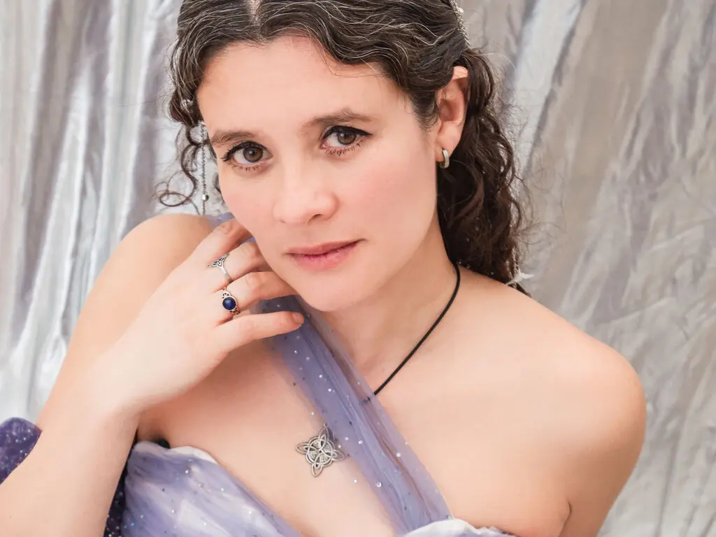 LiaMayerston wearing a purple dress and silver jewelry, posing with her hand near her face against a silver backdrop.