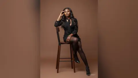 Elegant woman in a black long-sleeve dress and tights sitting on a wooden stool against a brown studio background.
