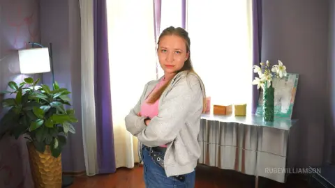 RubeWilliamson stands with her arms crossed, wearing a pink top, grey zip-up hoodie, and blue jeans in a room with a plant and a silver dresser.