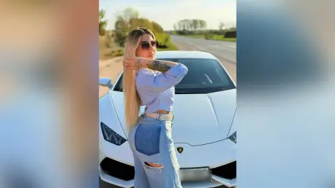 Stylish blonde woman in a blue striped shirt and denim jeans posing confidently by a white luxury sports car.