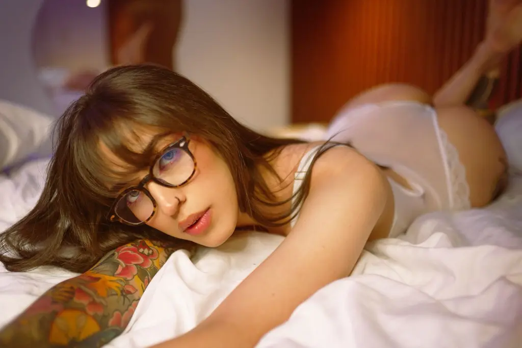 AlexNoir — Young woman with glasses in white lingerie lying on a bed in a cozy, intimate bedroom setting.