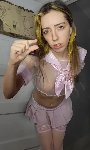 RixyRose wears a sheer pink schoolgirl outfit with a bow, showing her breasts and navel while posing in front of a wall.