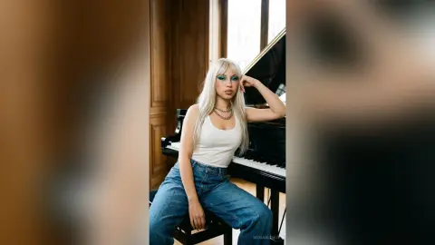Blonde woman in white tank top and blue jeans posing by a grand piano in a bright, elegant room.