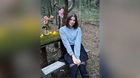 Young woman in a grey sweater and black skirt sitting by a candlelit table in a moody forest setting.