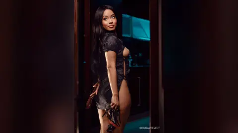 Glamorous woman in a black lace mini dress posing in a moody, dimly lit modern interior.