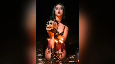 KimRoberth poses in black lingerie while holding a skull against her chest in a dark, candlelit setting.