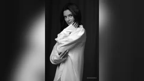 Elegant woman in a white oversized shirt posing against a dark studio background in a moody black and white portrait.