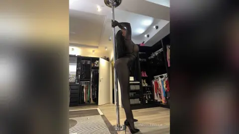 NicoleBella stands naked against a silver pole, wearing black sheer lingerie and black high heels while posing with her arms raised above her head.