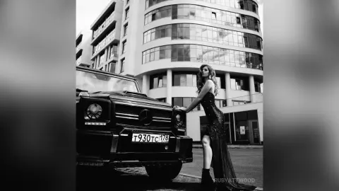 RubyAttwood poses next to a black SUV, wearing a long black lace dress that features a high slit showing her bare leg.